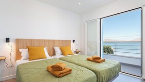 A modern hotel room with two beds, a wooden headboard, and a large balcony with a view of the sea.