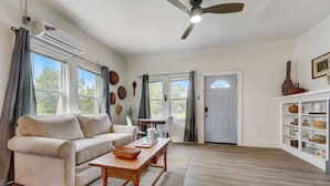 A living room with a sofa, coffee table, and a ceiling fan. There are windows, a door, and a shelf with decorative items. - Cozy Urban Getaway Near Paseo Arts 31A (Oklahoma City)