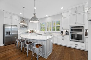 Una cocina moderna con armarios blancos, electrodomésticos de acero inoxidable, una isla grande con taburetes y vistas a la vegetación a través de las ventanas.