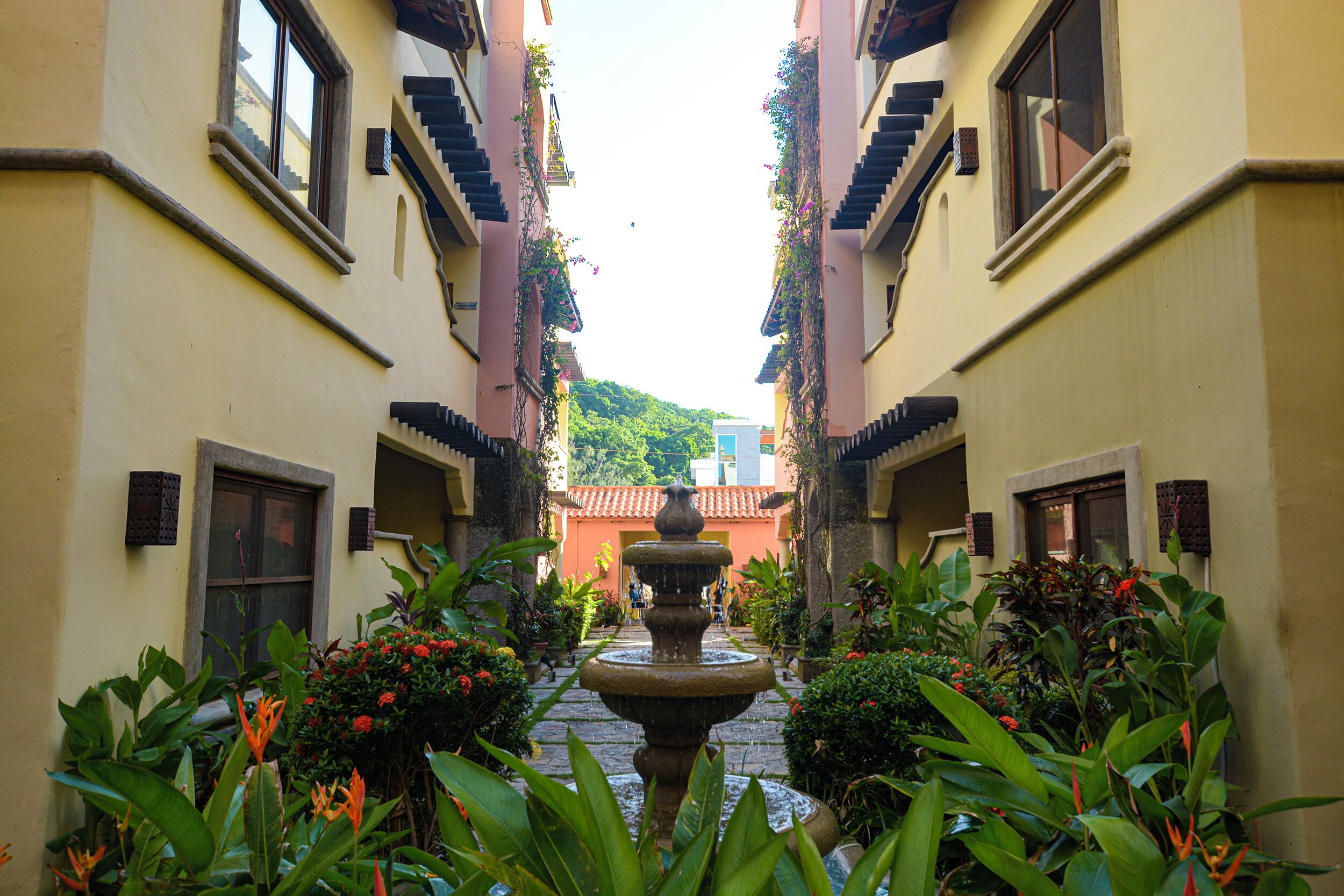 A narrow courtyard with a central fountain, surrounded by lush greenery and buildings with balconies.