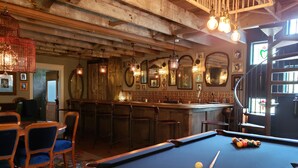 Un tavolo da biliardo con una stecca e palle, sgabelli da bar in legno, e un'area bar con luci a sospensione e specchi.