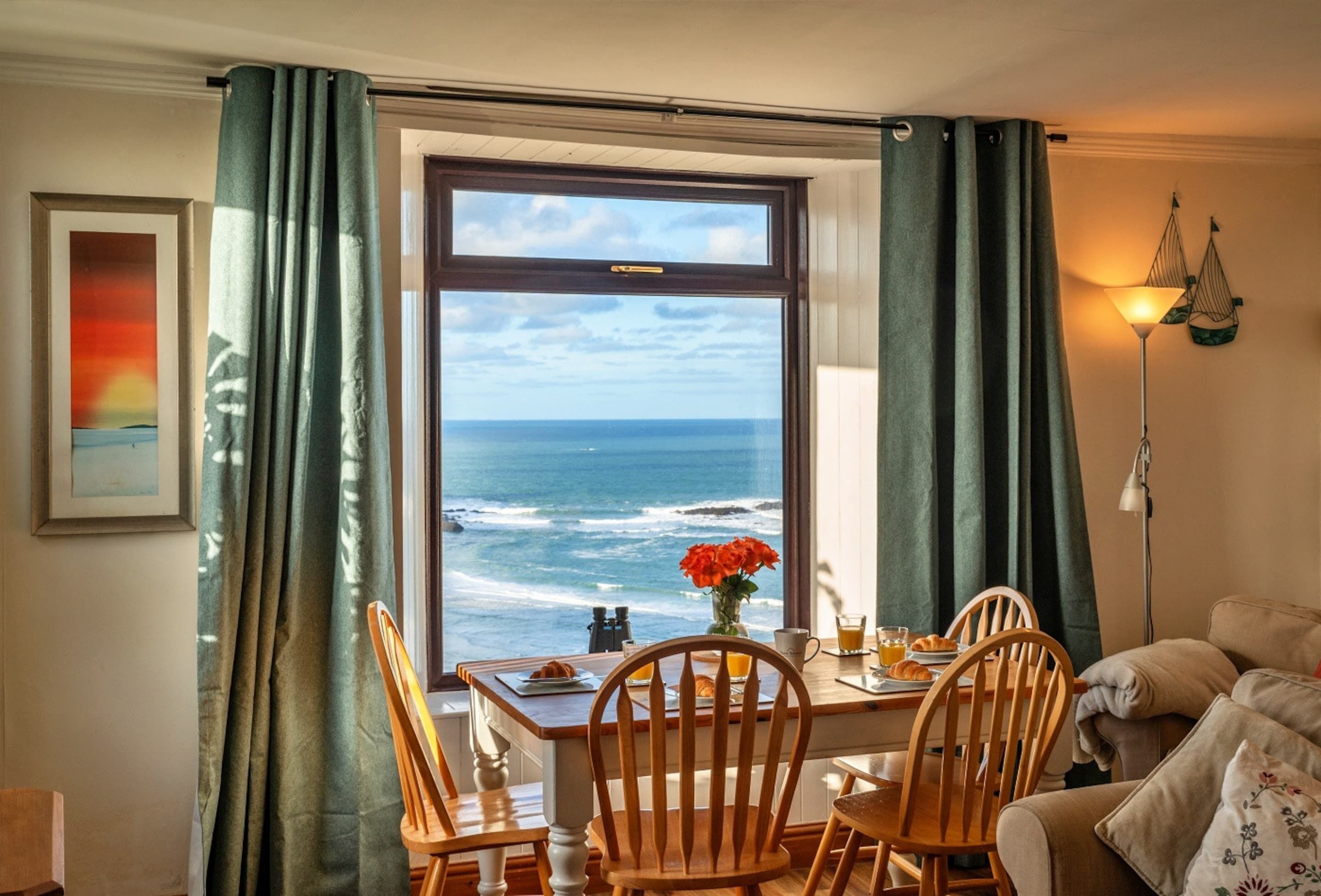 A dining area with a view of the ocean, a table with chairs, a vase with flowers, and a lamp on the side.