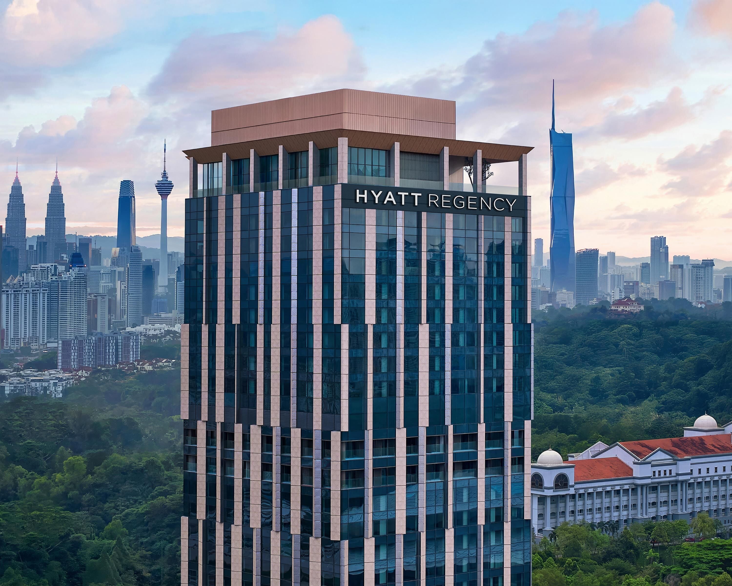 A modern high-rise hotel with a prominent name sign, surrounded by a dense urban skyline and lush greenery.