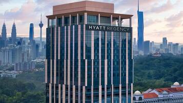 A modern high-rise hotel with a prominent name sign, surrounded by a dense urban skyline and lush greenery.