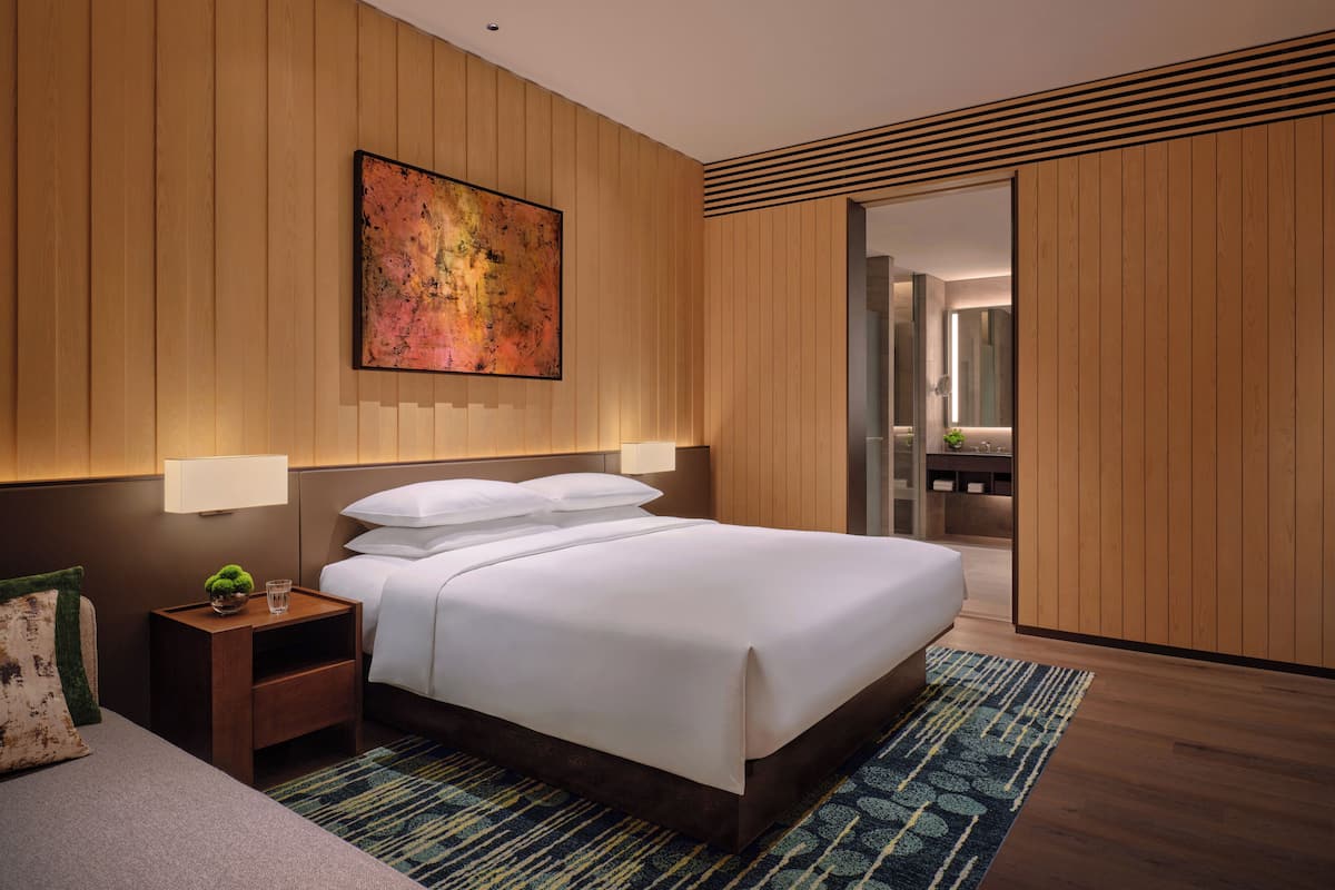 Una habitación de hotel moderna con una cama grande, paneles de madera, un cuadro y una mesita de noche con una lámpara.