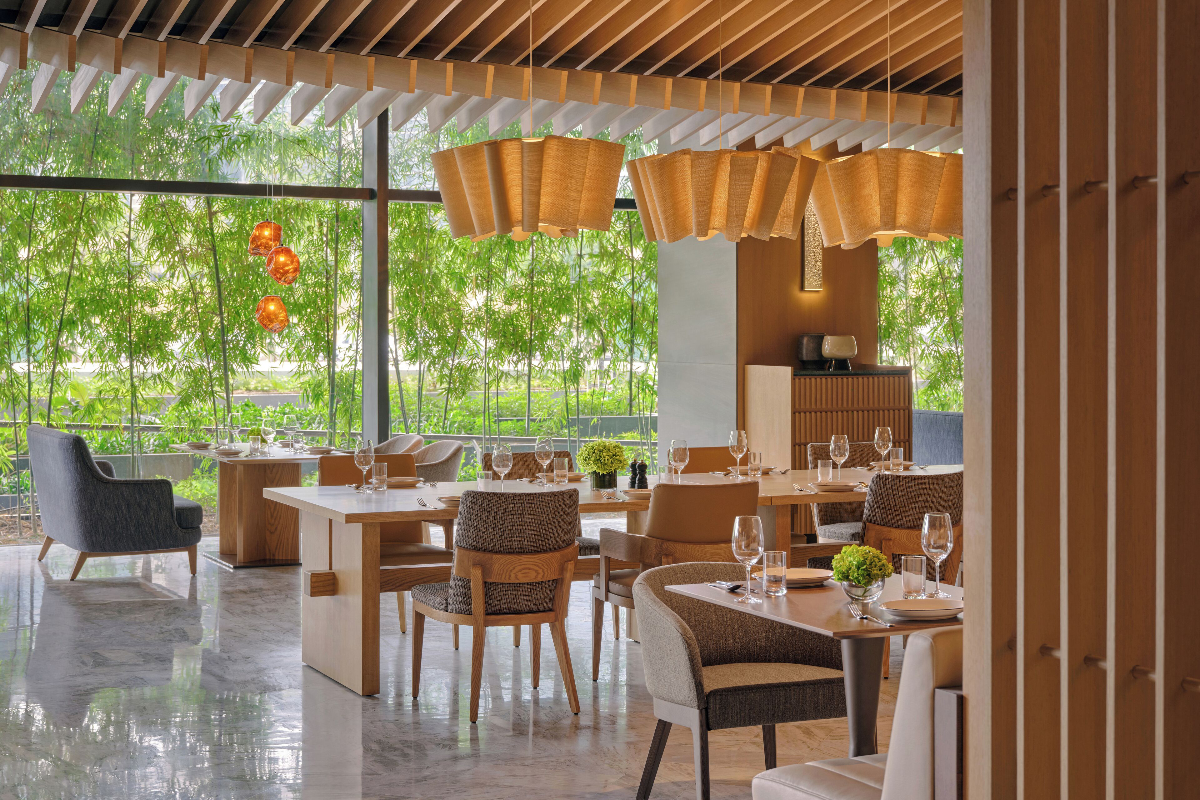 Un comedor moderno con mesas y sillas de madera, ventanas amplias con vistas a la vegetación y farolillos de papel colgantes.