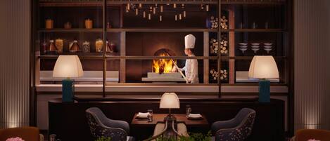 A restaurant with a chef at the counter, a fireplace, and a seating area with tables and chairs.