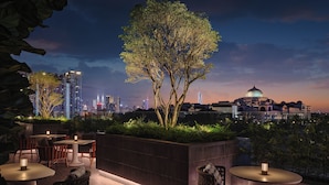 Un espacio al aire libre con mesas y sillas, un árbol grande y una vista panorámica de la ciudad de fondo.