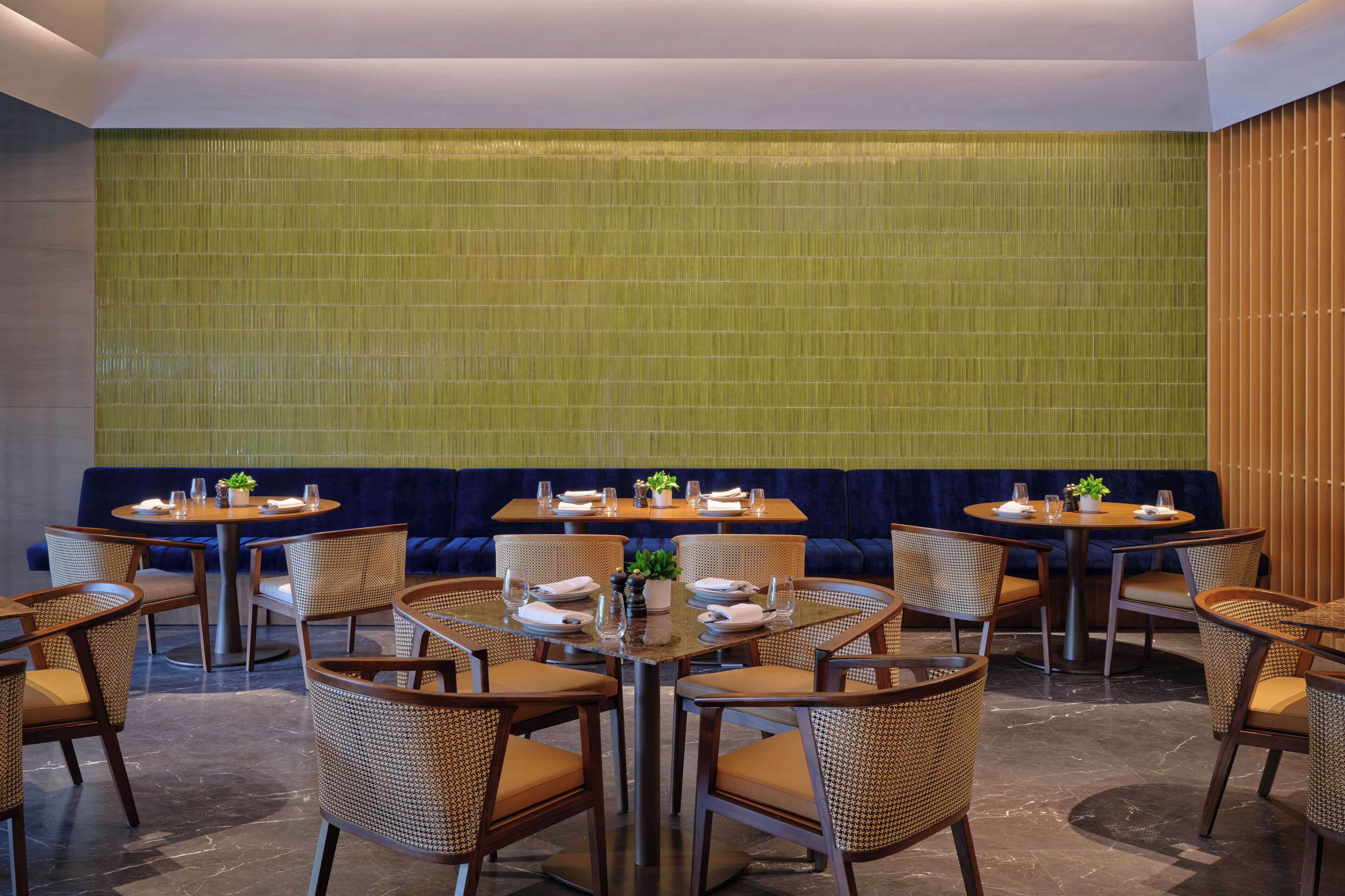 A restaurant with round tables and wicker chairs, a blue cushioned bench, and a green tiled wall.