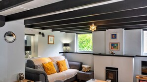 A cozy living room with a sofa, a small table, a fireplace, and a window with a view of greenery. - Cottage in Cornwall Near Porth Beach (Newquay)