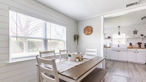 Une cuisine avec des meubles blancs, une table à manger en bois et un banc. On y trouve une fenêtre avec des stores, une horloge au mur et une plante suspendue.