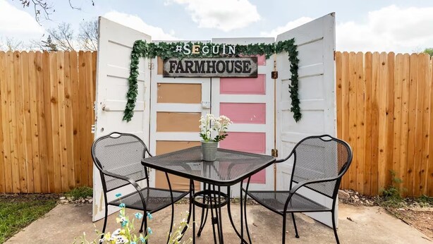 Une #FERME_SEGUIN avec une clôture en bois, une porte blanche ornée de panneaux roses et deux chaises en métal noir disposées autour d’une table sur laquelle se trouve un vase de fleurs.