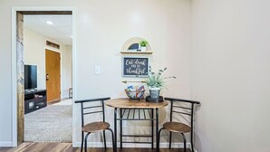 Um pequeno espaço de refeições com uma mesa e duas cadeiras, um letreiro na parede e uma planta sobre a mesa.