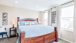 Quarto com cama de madeira, mesa de cabeceira, cadeira e janela com cortinas.