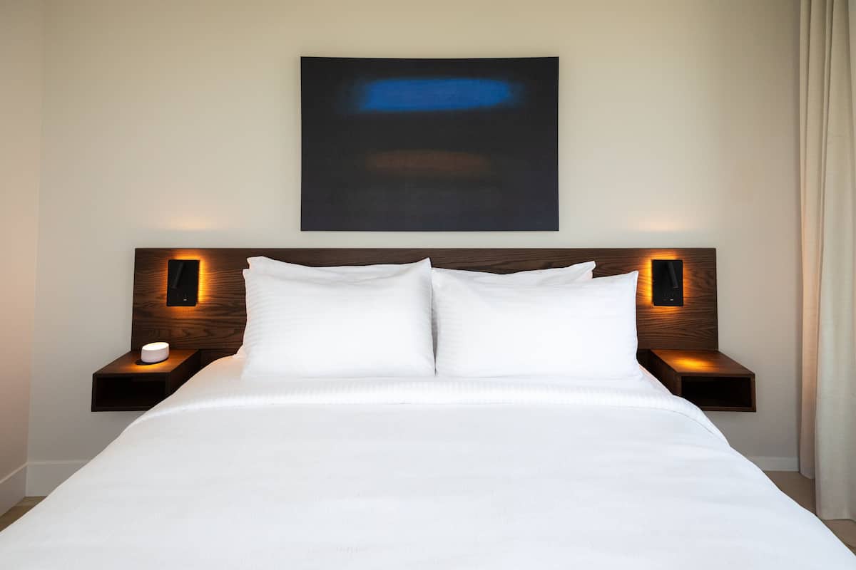 A neatly made bed with white linens, two bedside tables with illuminated wall sconces, and a large abstract painting above the bed.