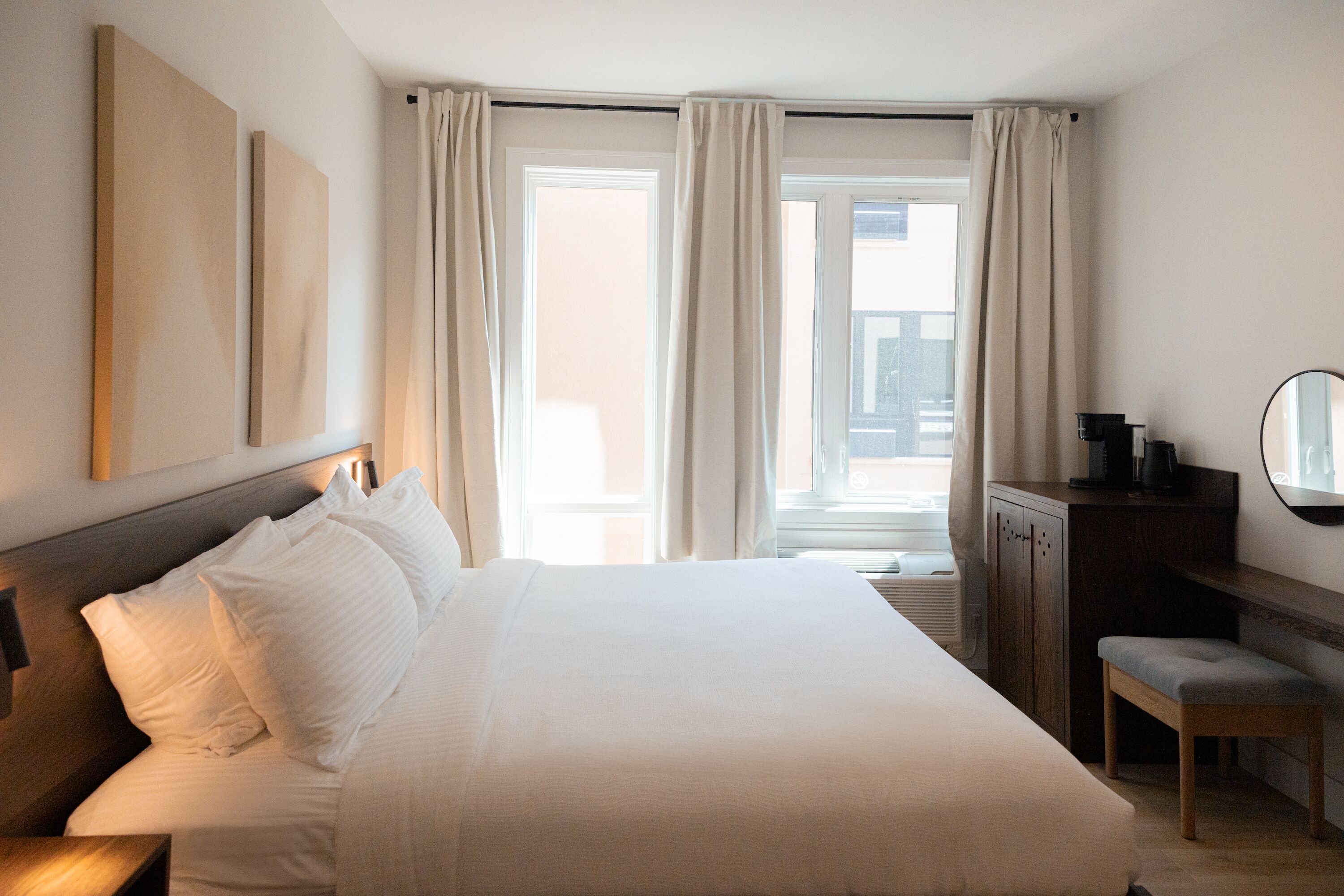 A hotel room with a large bed, a desk with a chair, a mirror, and a view of the city through the window.