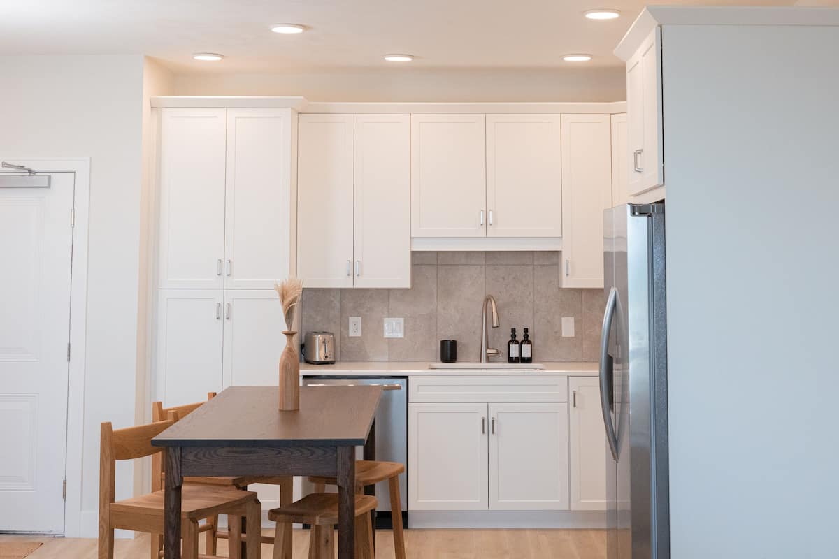 A modern kitchen with white cabinets, a wooden dining table, and a stainless steel refrigerator.