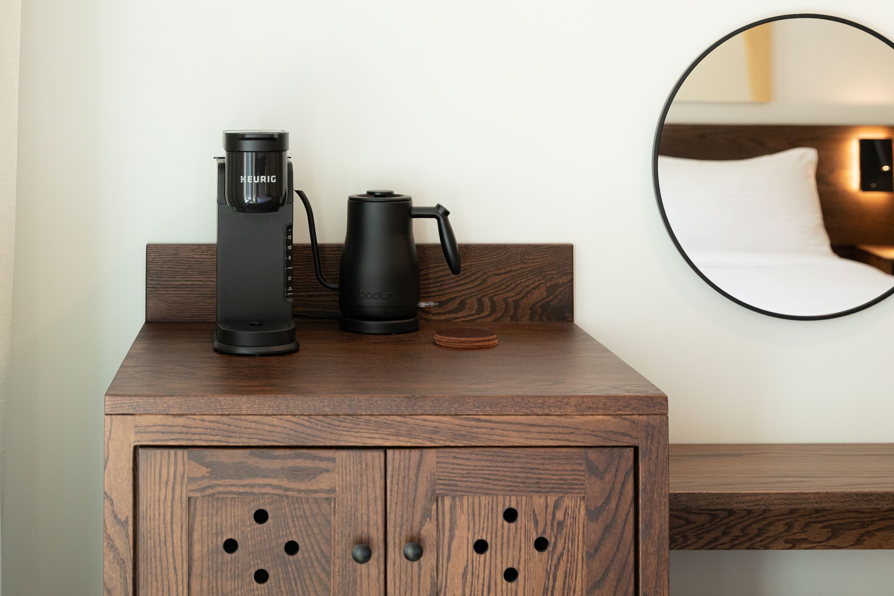 Une table de chevet en bois, avec une machine à café et une bouilloire, un miroir rond accroché au mur et un lit avec une tête de lit.