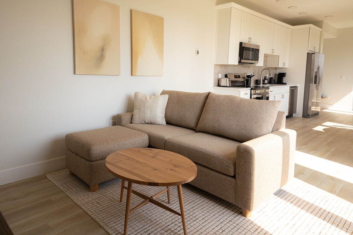 A modern living room with a beige sectional sofa, a wooden coffee table, and a kitchen in the background.