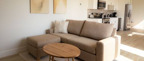 A modern living room with a beige sectional sofa, a wooden coffee table, and a kitchen in the background.