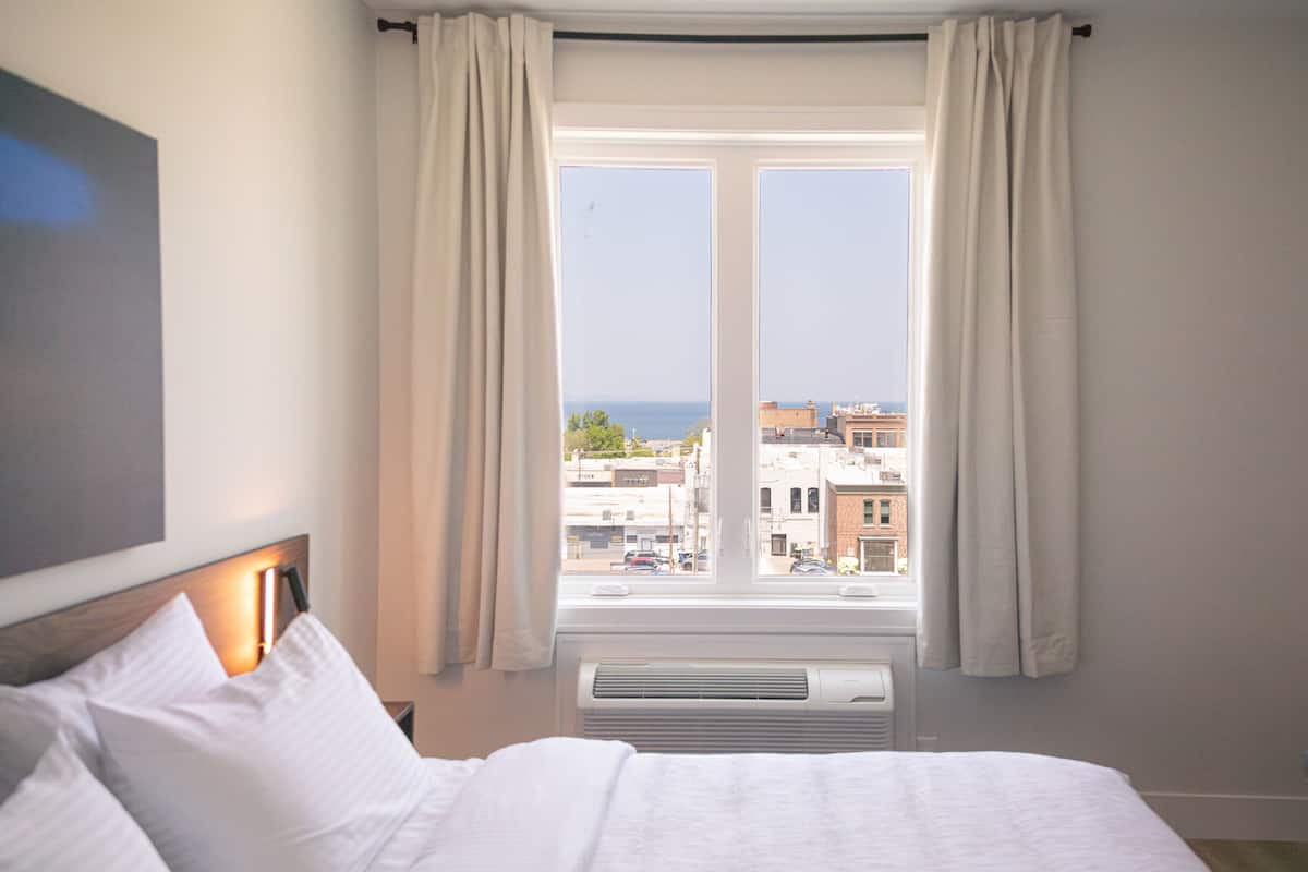 A hotel room with a large bed, a view of the city through a window, and a wall-mounted air conditioning unit.