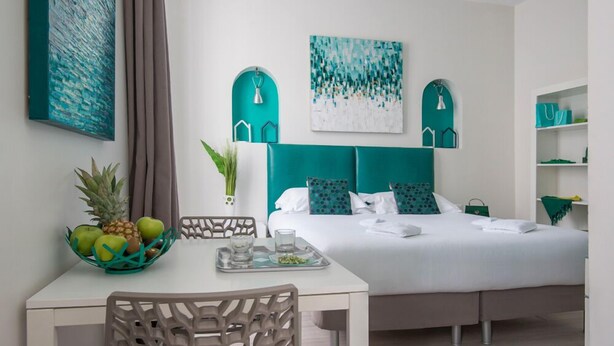 A modern hotel room with a bed, a dining table, and chairs. There are two wall-mounted shelves with decorative items and a painting above the bed. - O Carré Croisette (Cannes)