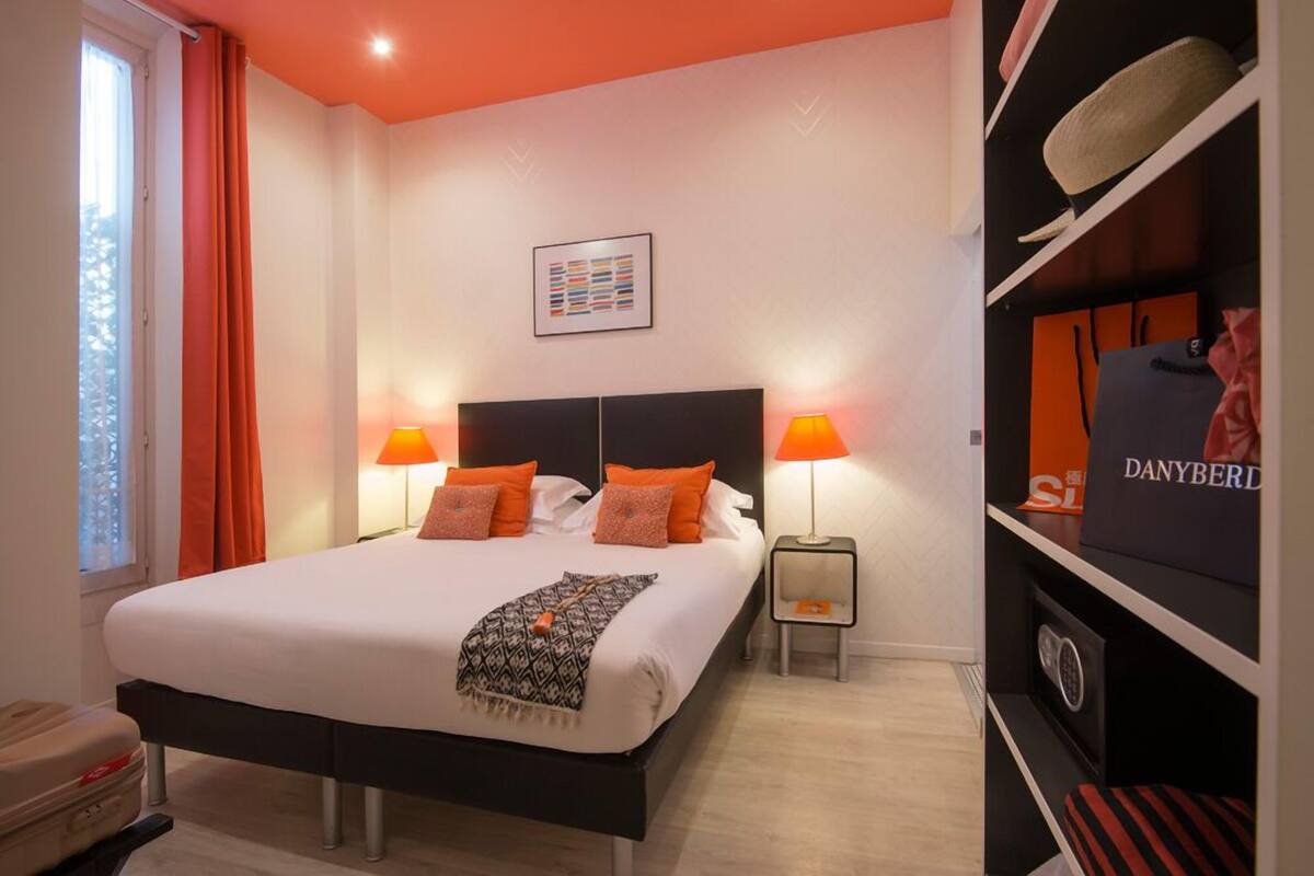 A hotel room with a large bed, two bedside lamps, a headboard, a small side table, a wardrobe, and a window with curtains.