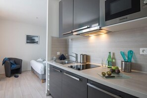 A modern kitchen with a microwave, sink, and stainless steel appliances. There is a bed and a chair in the adjacent room. - O Carré Croisette (Cannes)