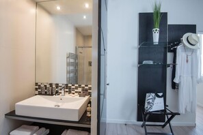 A modern bathroom with a large mirror, a white sink, and a black tiled backsplash. There is a glass shelf with a potted plant and a hat. - O Carré Croisette (Cannes)