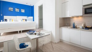 A modern hotel room with a white bed, a small dining area with a white table and chairs, a kitchen with white cabinets and a microwave, and a blue accent wall. - O Carré Croisette (Cannes)