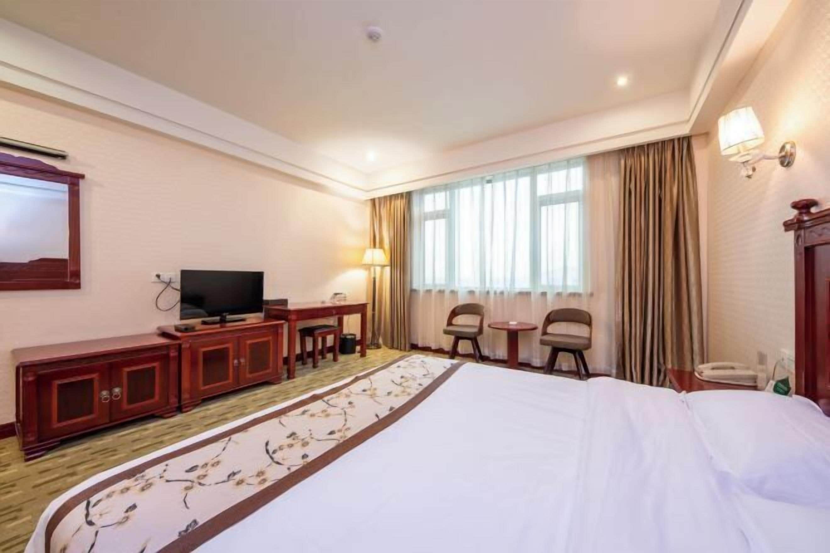 Une chambre d’hôtel comprenant un lit, une télévision, un bureau avec une chaise, une petite table et une fenêtre avec des rideaux.