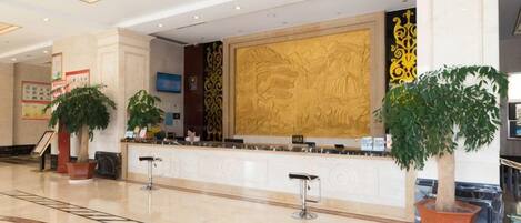 A hotel lobby with a reception desk, potted plants, and a large decorative wall.