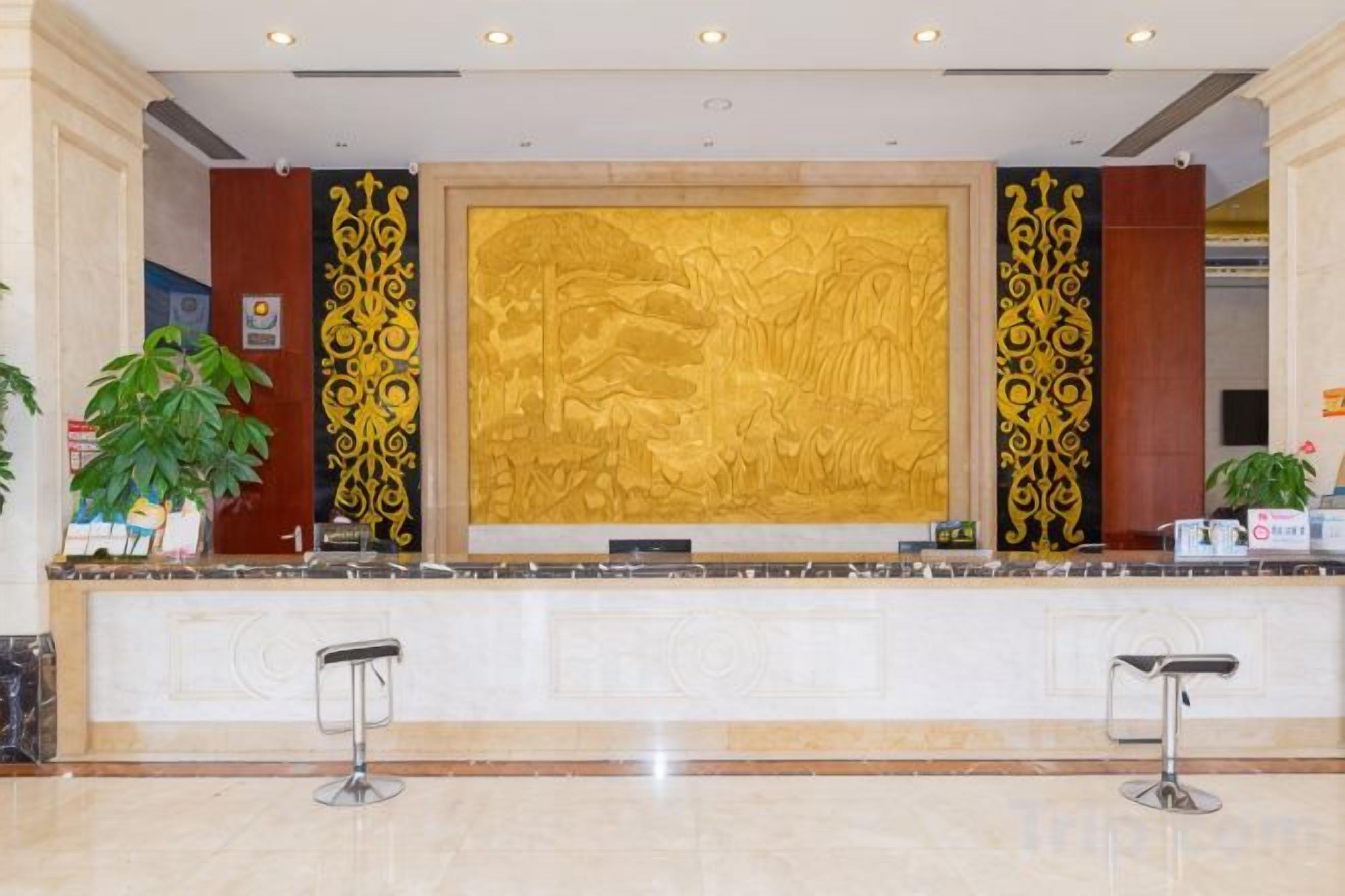 A reception desk with a large golden relief sculpture, two bar stools, and potted plants.