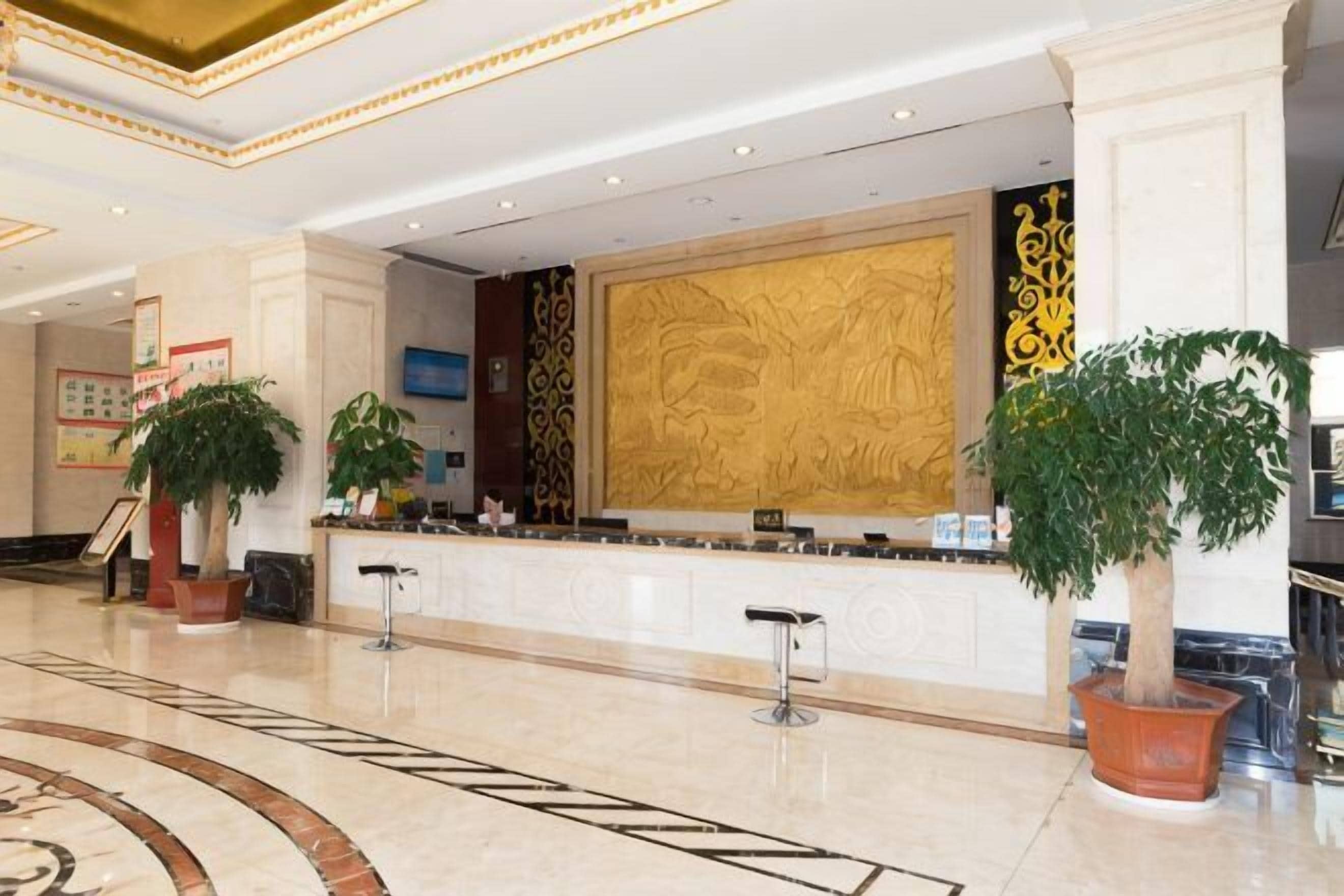A hotel lobby with a reception desk, potted plants, a large decorative wall, and a marble floor.