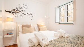 Een slaapkamer met een bed, kussens, een nachtkastje met boeken, een lamp, een wanddecoratie van een boom en een raam met uitzicht.