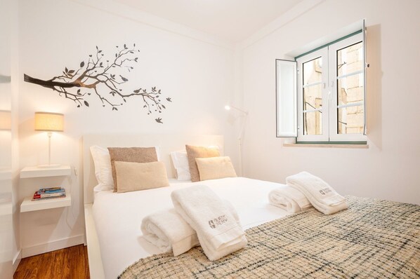 Een slaapkamer met een bed, kussens, een nachtkastje met boeken, een lamp, een wanddecoratie van een boom en een raam met uitzicht.