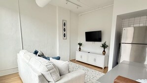 A modern living room with a sofa, a white cabinet, a flat-screen TV, and a kitchen area with a refrigerator.