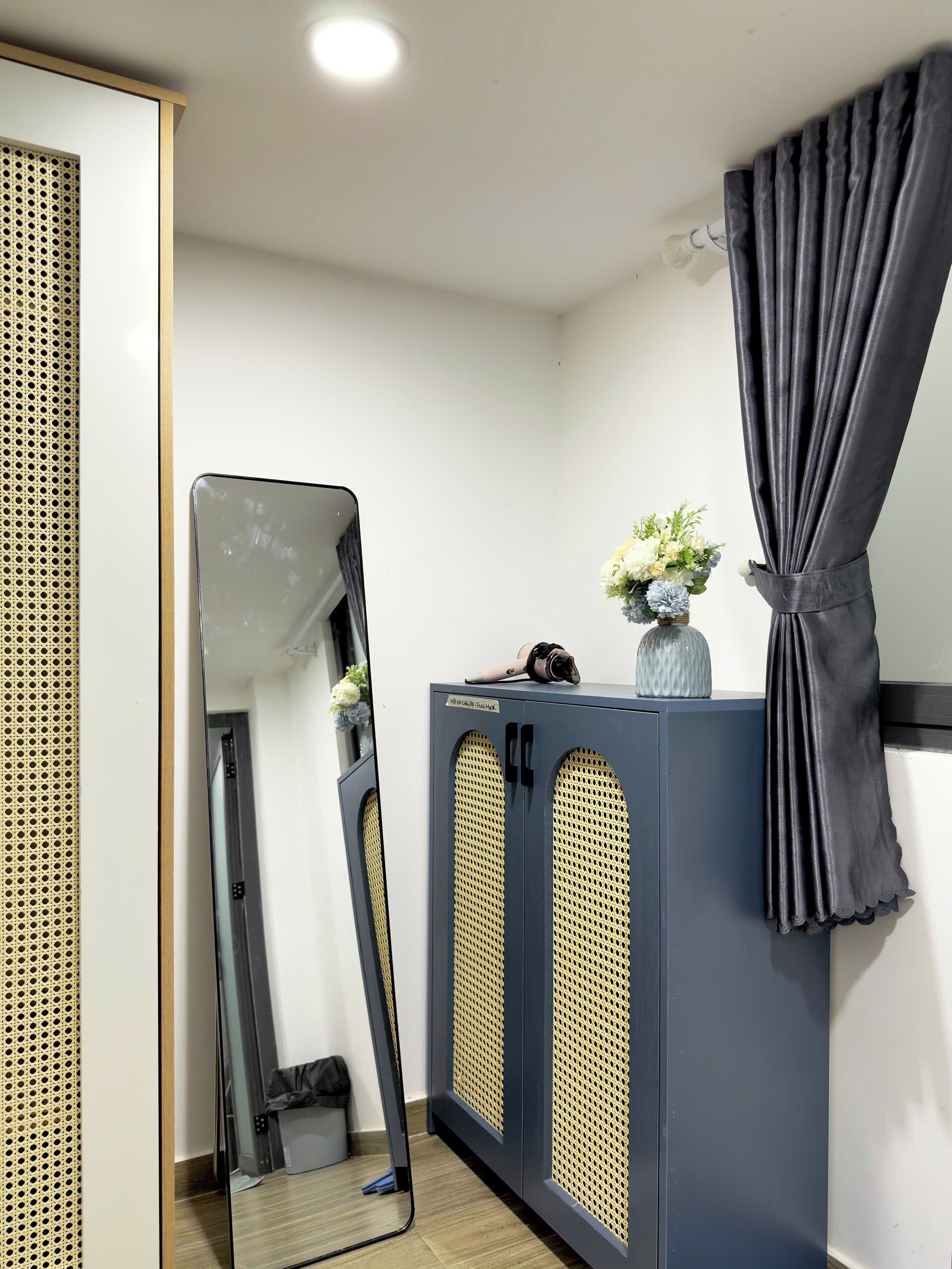 Une pièce avec un grand miroir, une armoire dotée de panneaux perforés, un rideau foncé et un vase de fleurs posé sur une commode.