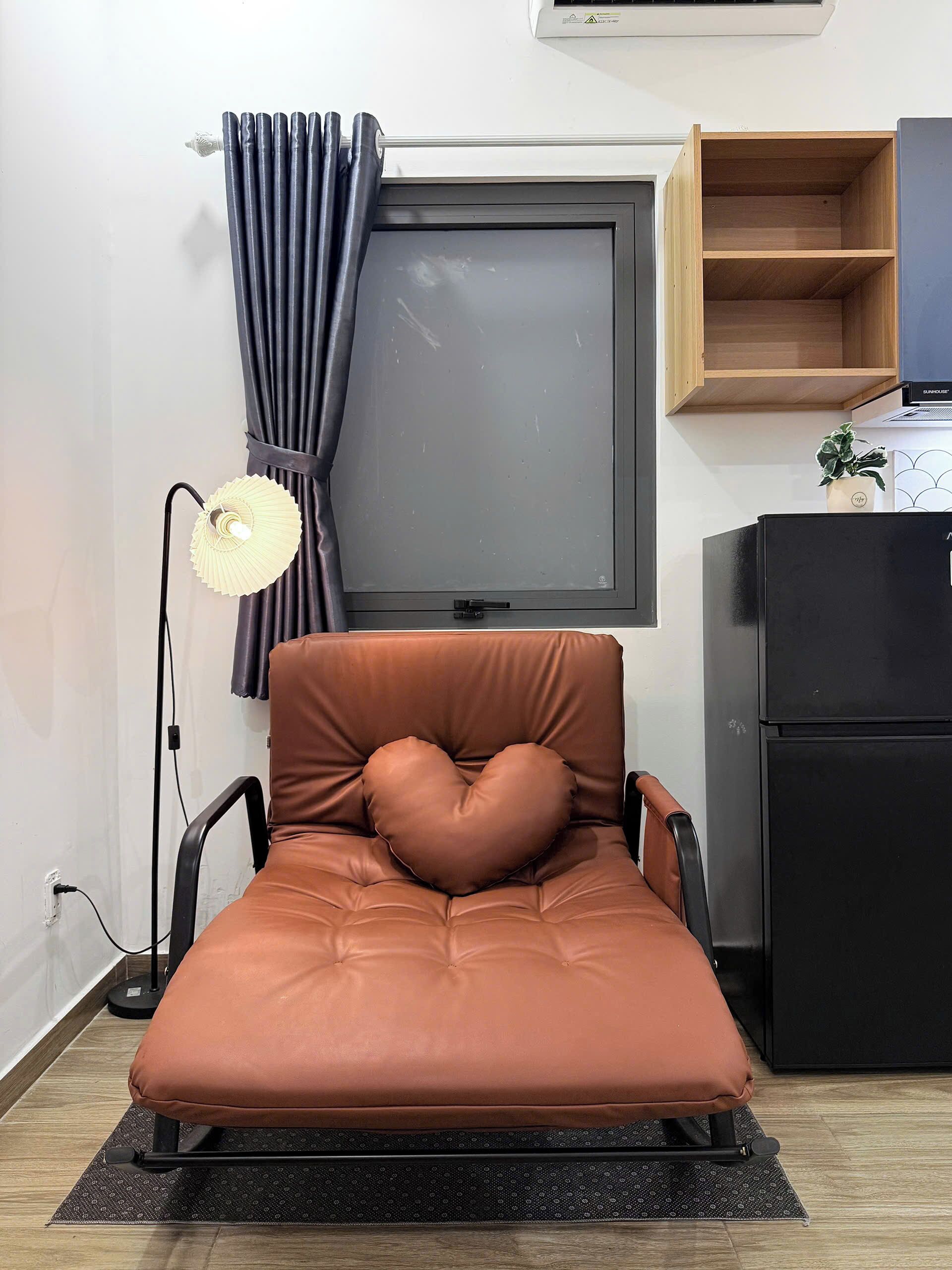 A single bed with a heart-shaped pillow, a standing lamp, a window with curtains, a small wooden shelf, and a black refrigerator.