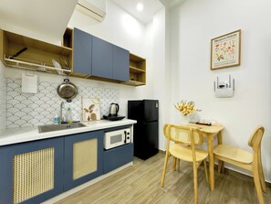 Una cocina compacta con microondas, hervidor y un refrigerador negro. Hay una zona de comedor con dos sillas y una mesa, y un cuadro en la pared.