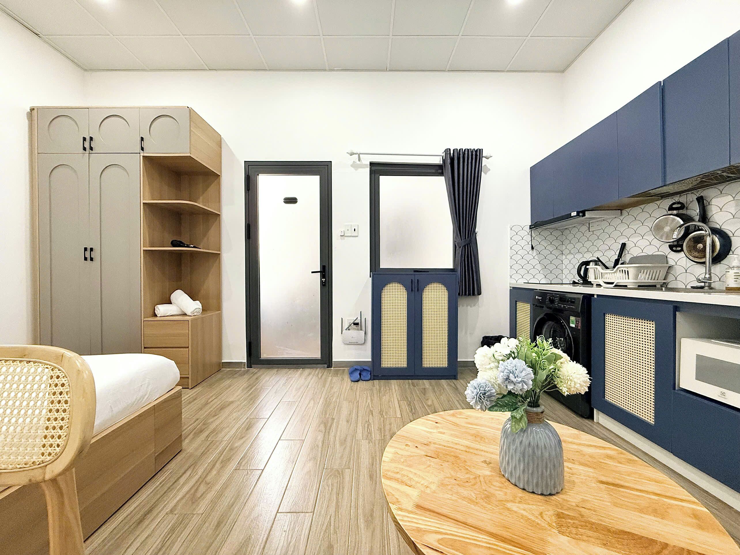 A compact hotel room with a bed, a small table with a vase, a wardrobe, a door, a kitchen area with a washing machine, and a window with curtains.