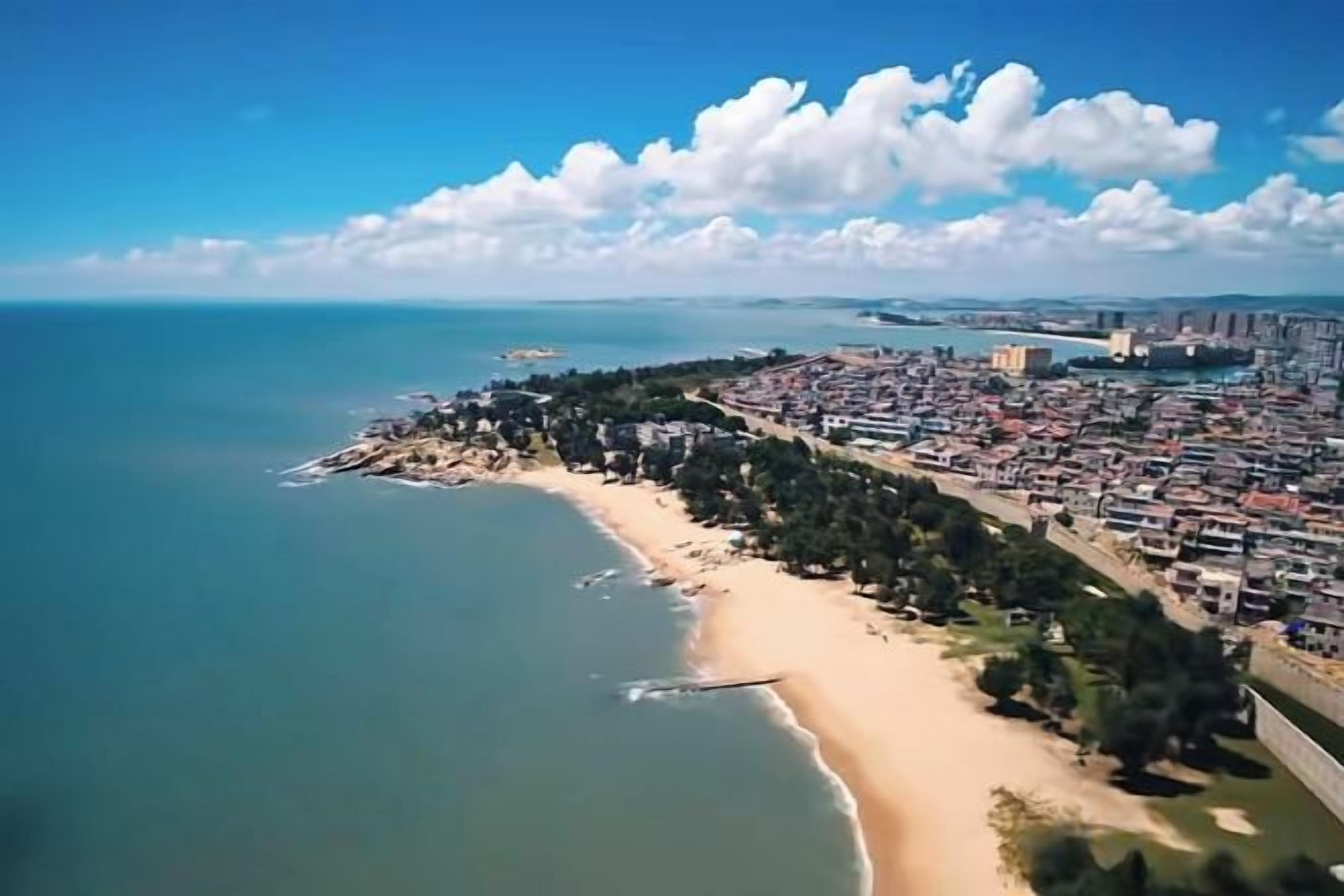 An aerial view of a coastal city with a beach, dense urban area, and a clear sky.