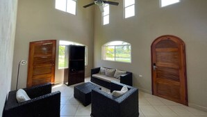 A spacious living area with a high ceiling, a ceiling fan, and a large window providing natural light. There is a sofa and two wicker chairs, and a wooden door is visible in the background. - Casa Inge: 3BR Golf-Front Villa at Gran Pacifica Resort. Pet Friendly! (Gran Pacifica Resort)