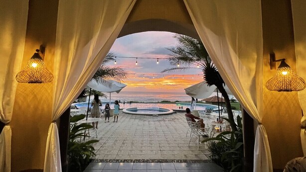 An outdoor seating area with a view of the sunset over the water, featuring a pool and palm trees. - 2BR Eco-Tiny Home Near the Beach by Veritas Pacifica • T4 (Gran Pacifica Resort)