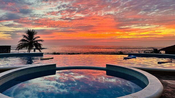 Ein Pool mit Blick auf einen Sonnenuntergang, eine Palme und ein Strand im Hintergrund.