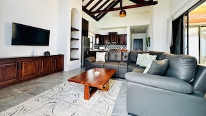 Ein Wohnzimmer mit Couchtisch aus Holz, einem grauen Sofa, einem Flachbildfernseher und einer Küche im Hintergrund.
