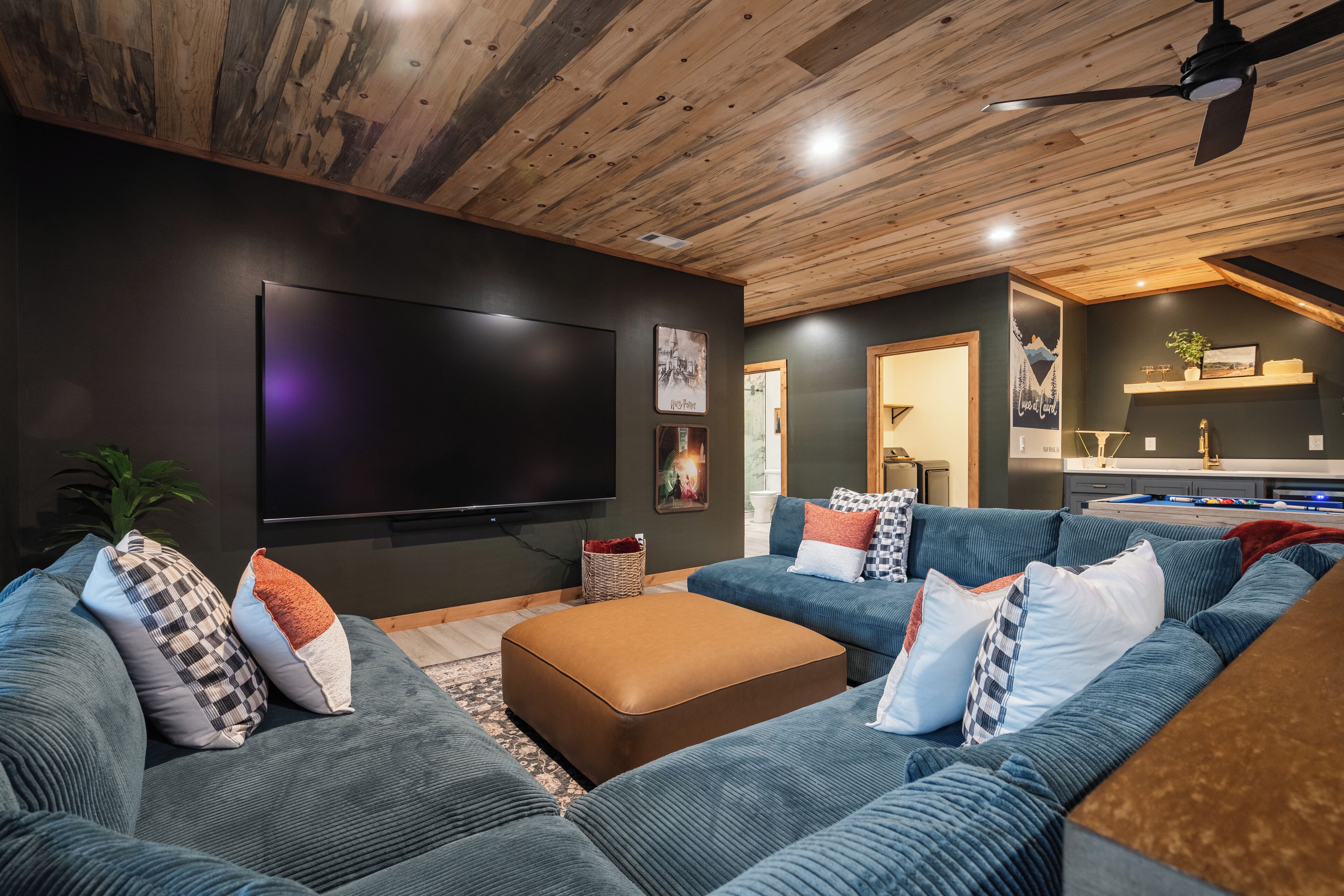 A modern living room with a large flat-screen TV, dark walls, and a wooden ceiling.