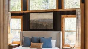 A bedroom with a large bed, a headboard, a nightstand, a lamp, and a view of trees through the windows. - New Luxe at Laurel - Putting Green in Blue Ridge (Blue Ridge)