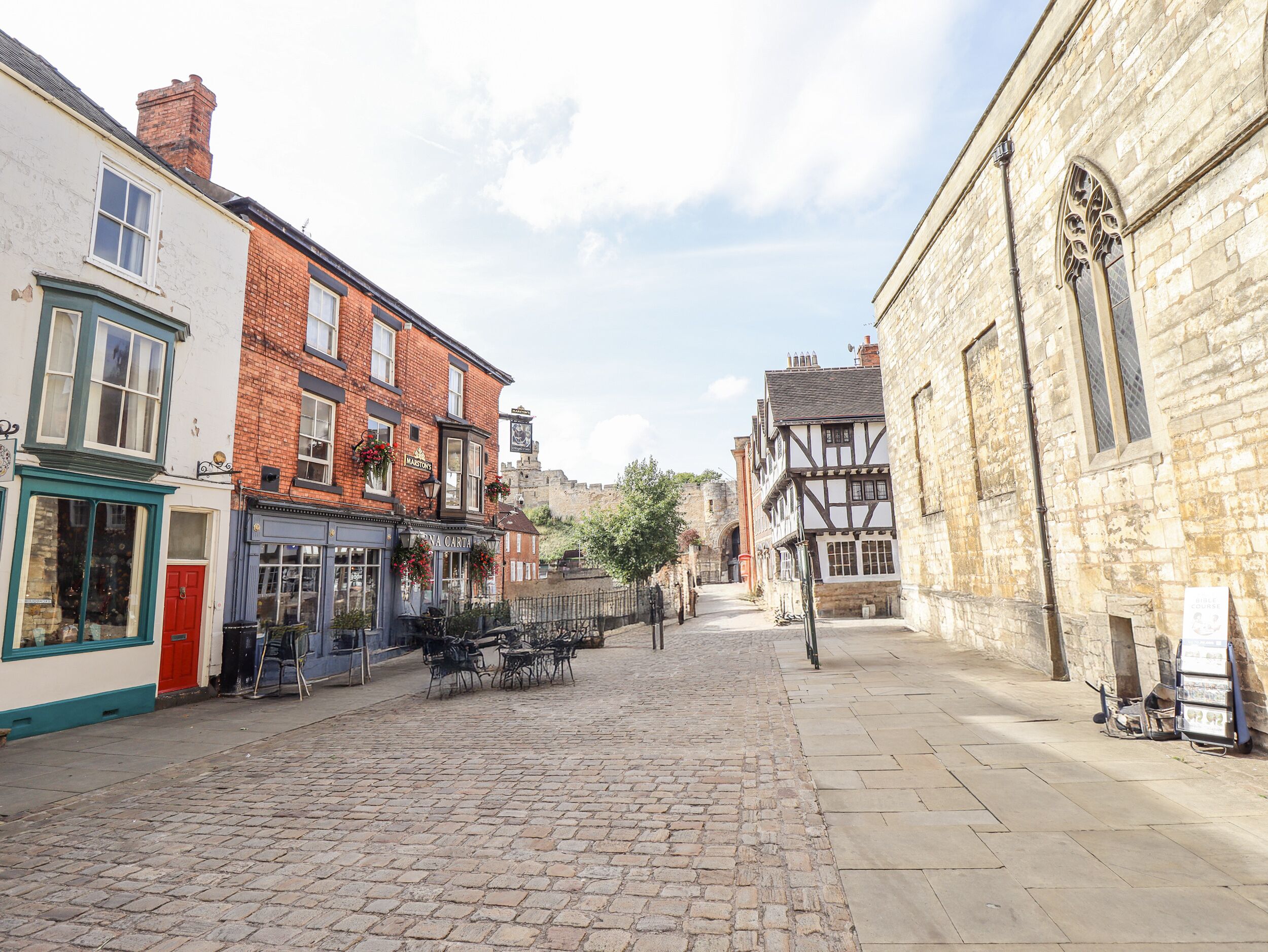 A cobblestone street lined with historic buildings, including a pub with outdoor seating and a sign reading "MASTONS."
