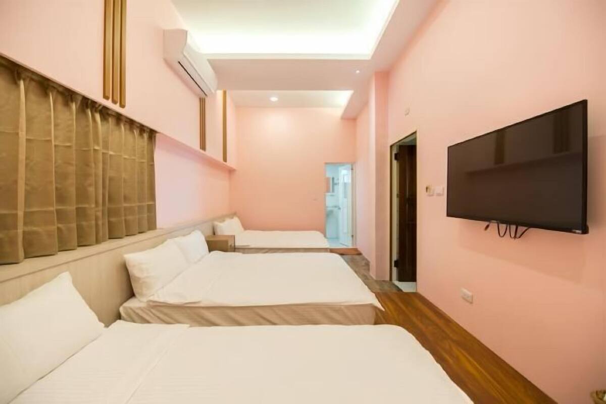 A hotel room with three beds, a flat-screen TV mounted on the wall, and a window with curtains.